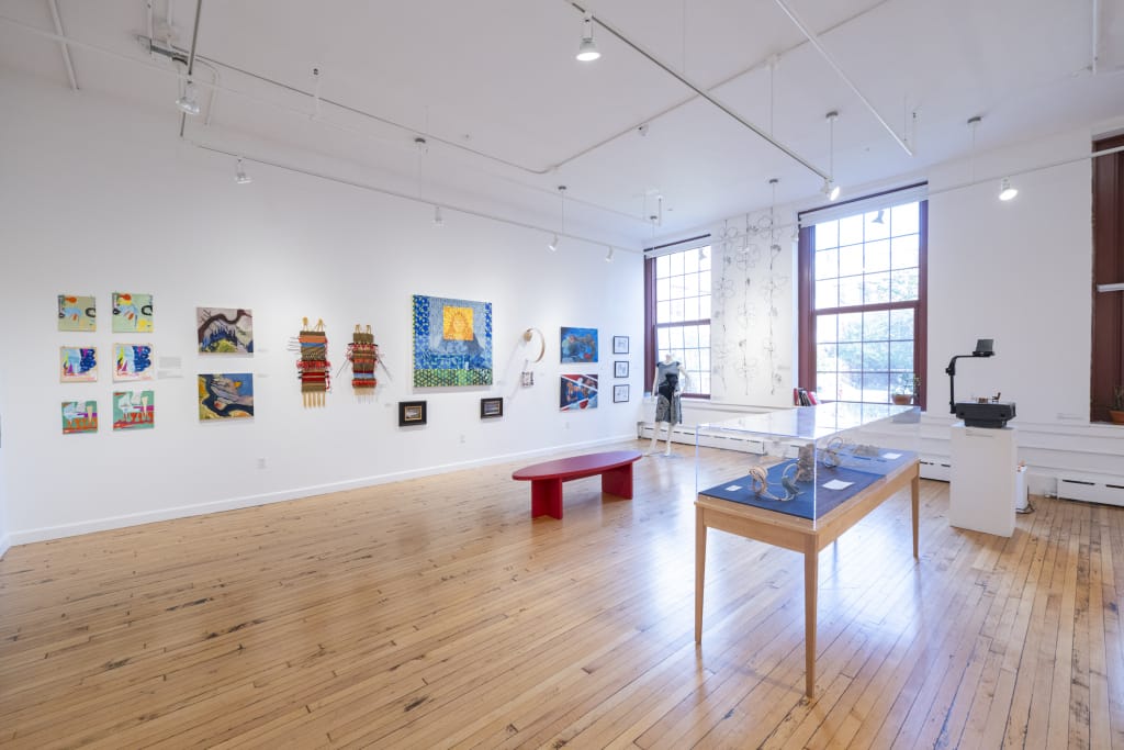 A gallery space with artwork on white walls, some tables, and light streaming in from the outdoors