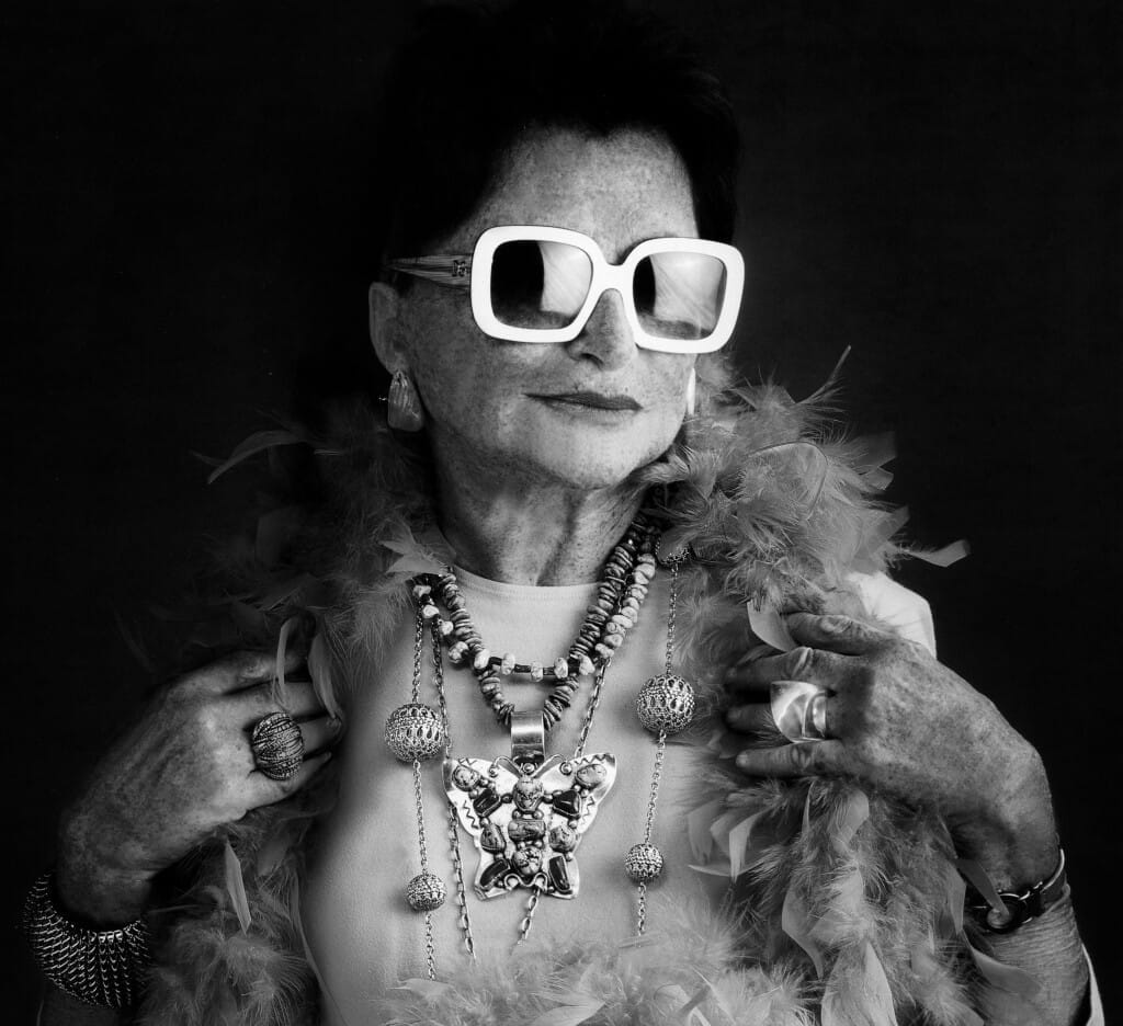 A stylish older woman with short hair wears large white sunglasses, statement jewelry, and a feather boa. She poses confidently against a dark background, enhancing her elegant and bold appearance. The image is in black and white.
