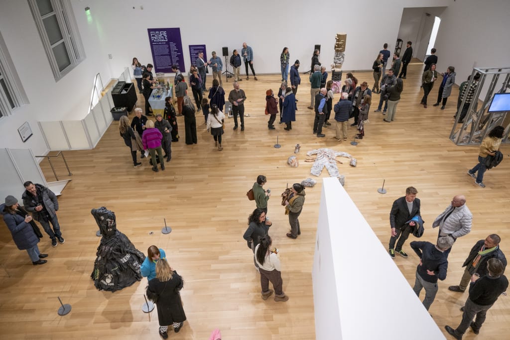 A spacious art gallery at MassArt with wooden floors, where numerous visitors observe various sculptures and artworks. The room is bustling with people conversing and viewing displays on pedestals under bright lighting, reminiscent of a vibrant art museum atmosphere.
