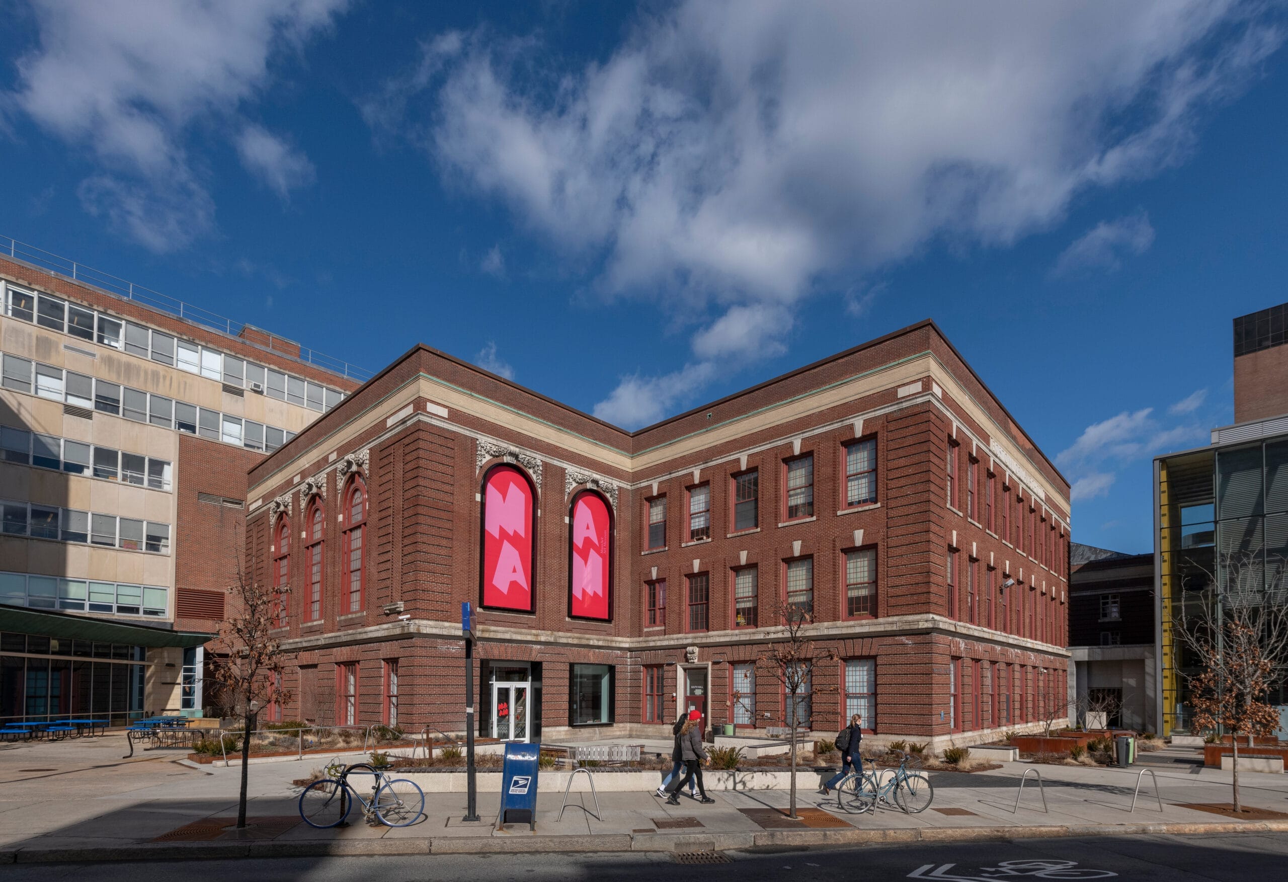 A large, three-story brick building with tall windows, displaying red banners. People walk on the sidewalk in front, and bicycles are parked nearby. The sky is partly cloudy, and the scene has a campus-like feel.