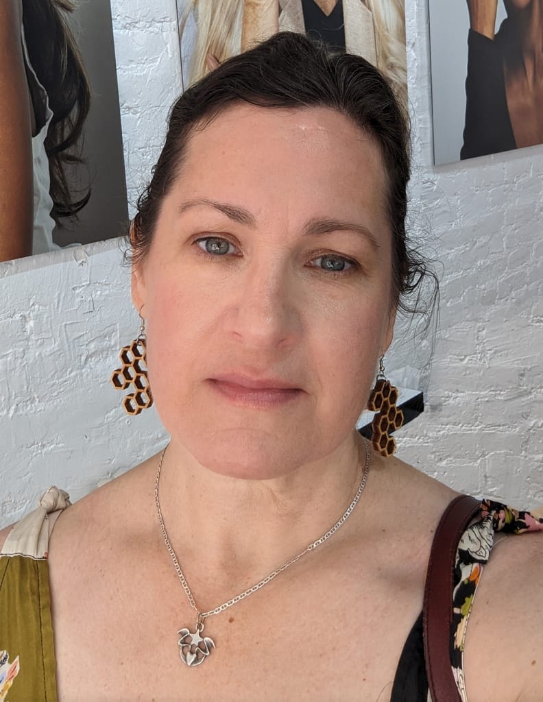 Woman with brown hair and leopard print earrings, wearing a necklace with a leaf pendant. She is standing in front of a white brick wall, partially visible portraits are displayed above.