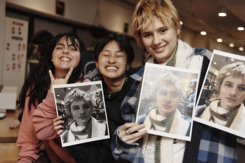 Three people, possibly from a student organization, smile indoors while holding printed photos. Two hold images of someone with similar features. They appear joyful and relaxed in a casual setting, enhanced by warm lighting in the background.