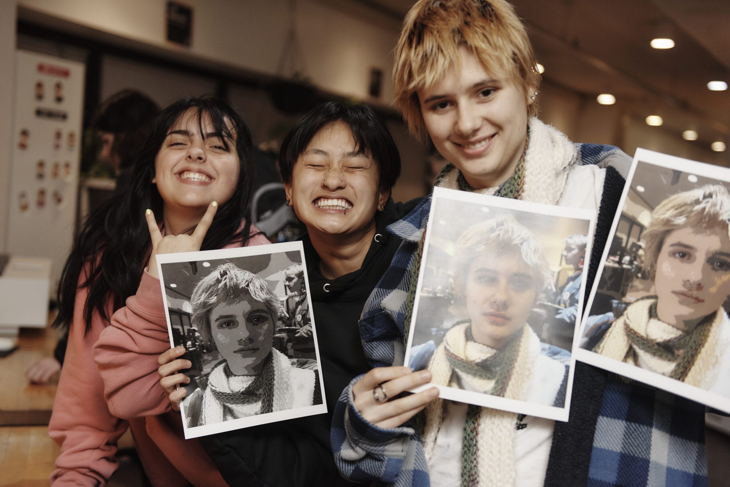 Three people, possibly from a student organization, smile indoors while holding printed photos. Two hold images of someone with similar features. They appear joyful and relaxed in a casual setting, enhanced by warm lighting in the background.