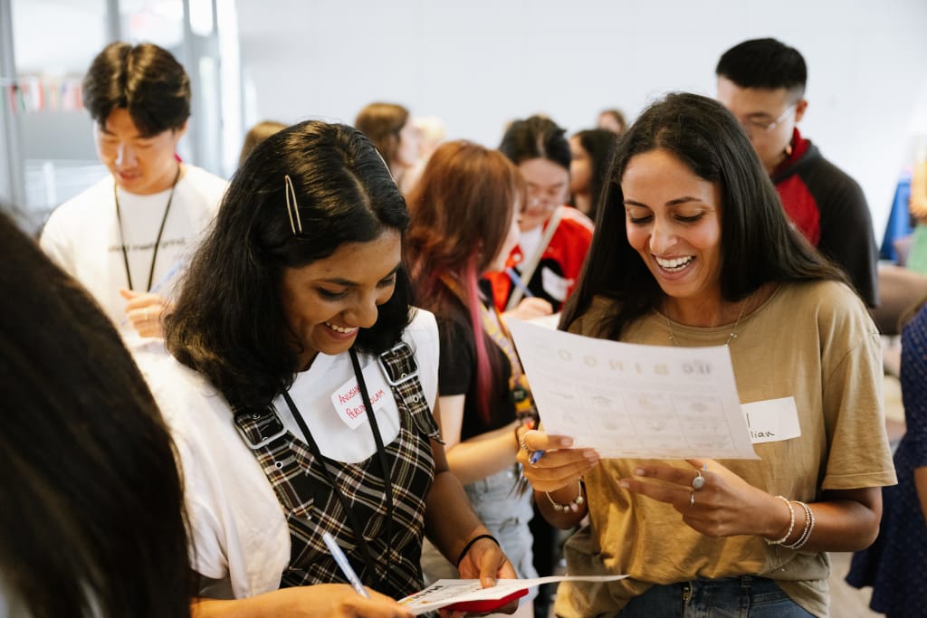 A group of diverse individuals are gathered in a room, representing various student organizations. Two women in the foreground smile while reading papers. Others, wearing casual clothes and name tags, stand and interact in the background, fostering a lively atmosphere of collaboration.