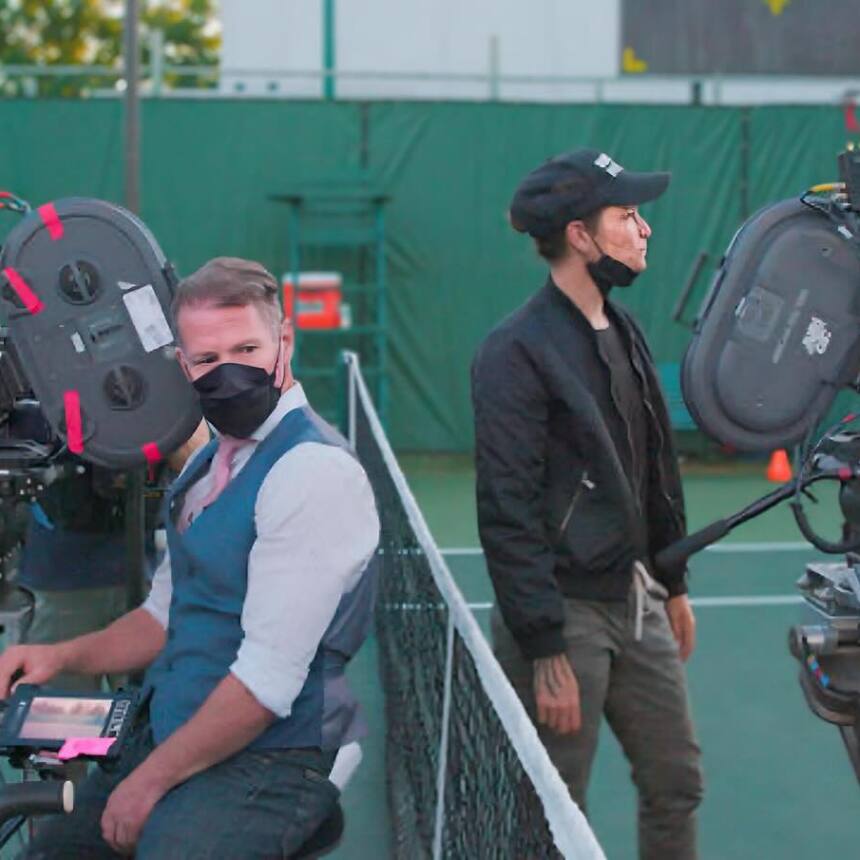 Two masked crew members operate film cameras on a tennis court set.