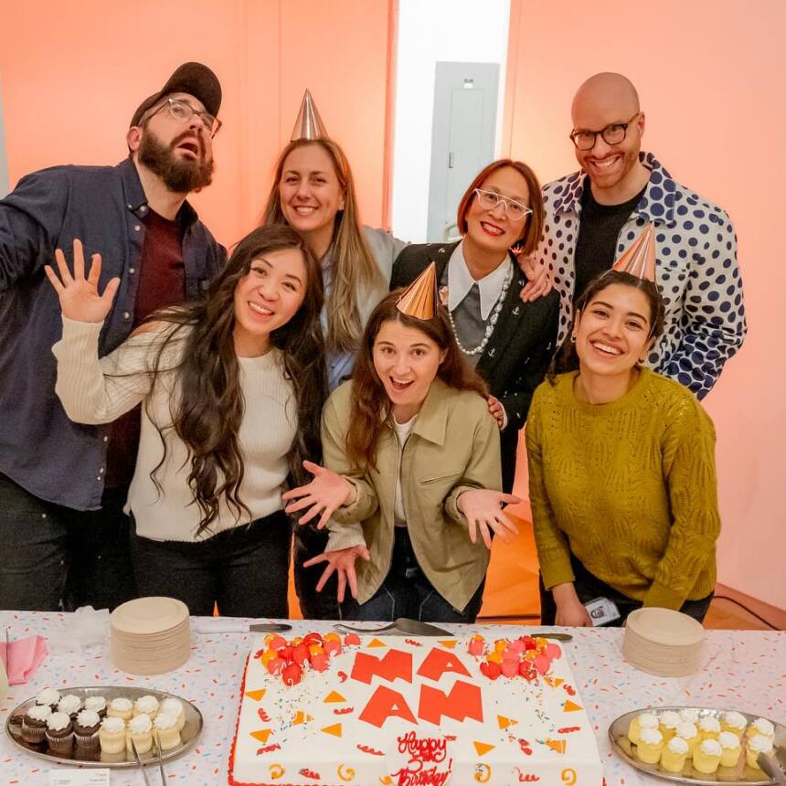 A group of seven people smiling and posing behind a table adorned with a beautifully decorated cake at the MassArt Art Museum (MAAM). Some people don festive party hats. The spread includes cupcakes and plates and the backdrop glows with a light pink-orange hue.