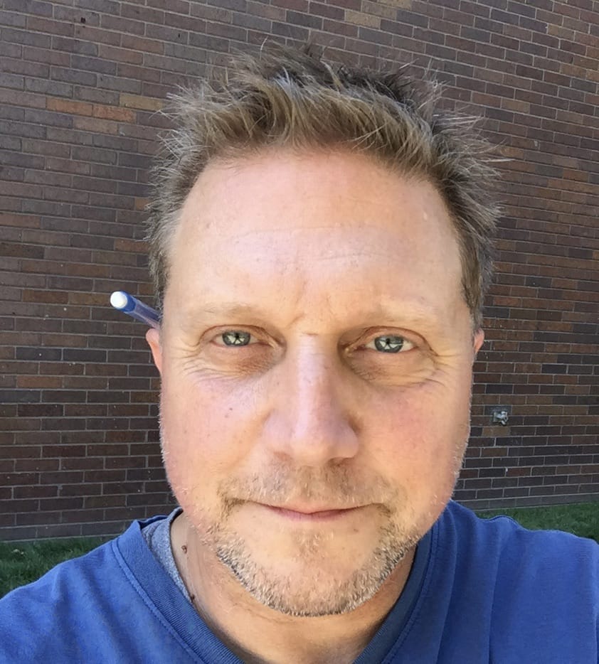 A middle-aged man with short, tousled hair and light facial stubble is looking directly at the camera. Hes wearing a blue shirt and has a pen tucked behind his ear. The background is a brick wall with a patch of green grass.