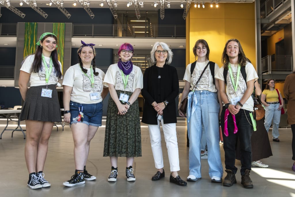 A group of six people stands indoors at MassArt, smiling at the camera. Five are in casual festival attire with lanyards, while the one in the center wears a black blazer and white pants. The modern architectural space highlights their vibrant energy and creativity.