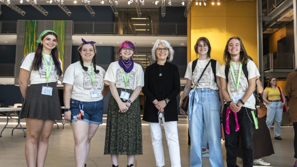 A group of six people stands in a spacious indoor setting at MassArt. They are casually dressed, with five wearing graphic tees and accessories like bandanas. One sports black and white clothing. The background is marked by tall windows and tables, adding an artistic touch to the scene.
