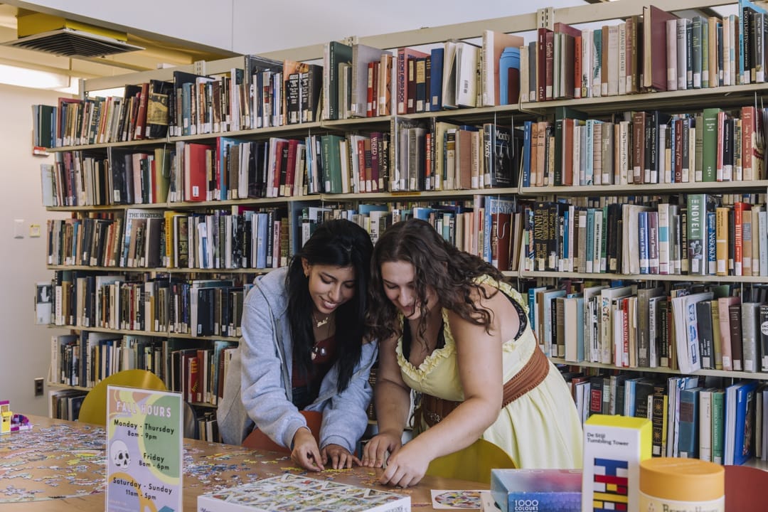 Two women are focused on assembling a puzzle on a table in a library, surrounded by bookshelves filled with books. The bright and inviting scene promotes an engaging atmosphere that nurtures well-being and curiosity.