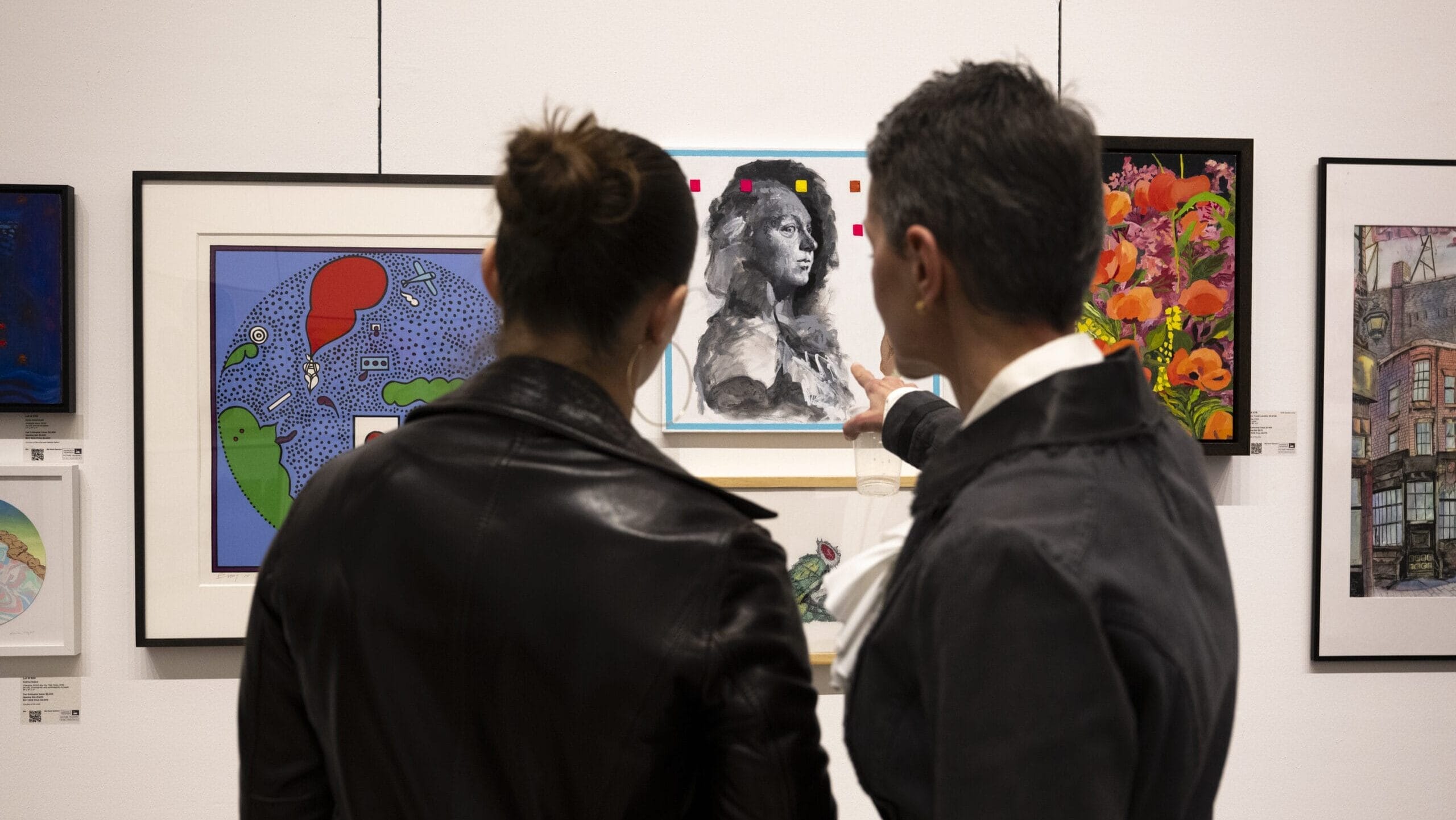 Two people in jackets stand in front of an art display. One points towards a black and white portrait piece. Colorful artwork surrounds them on the wall.
