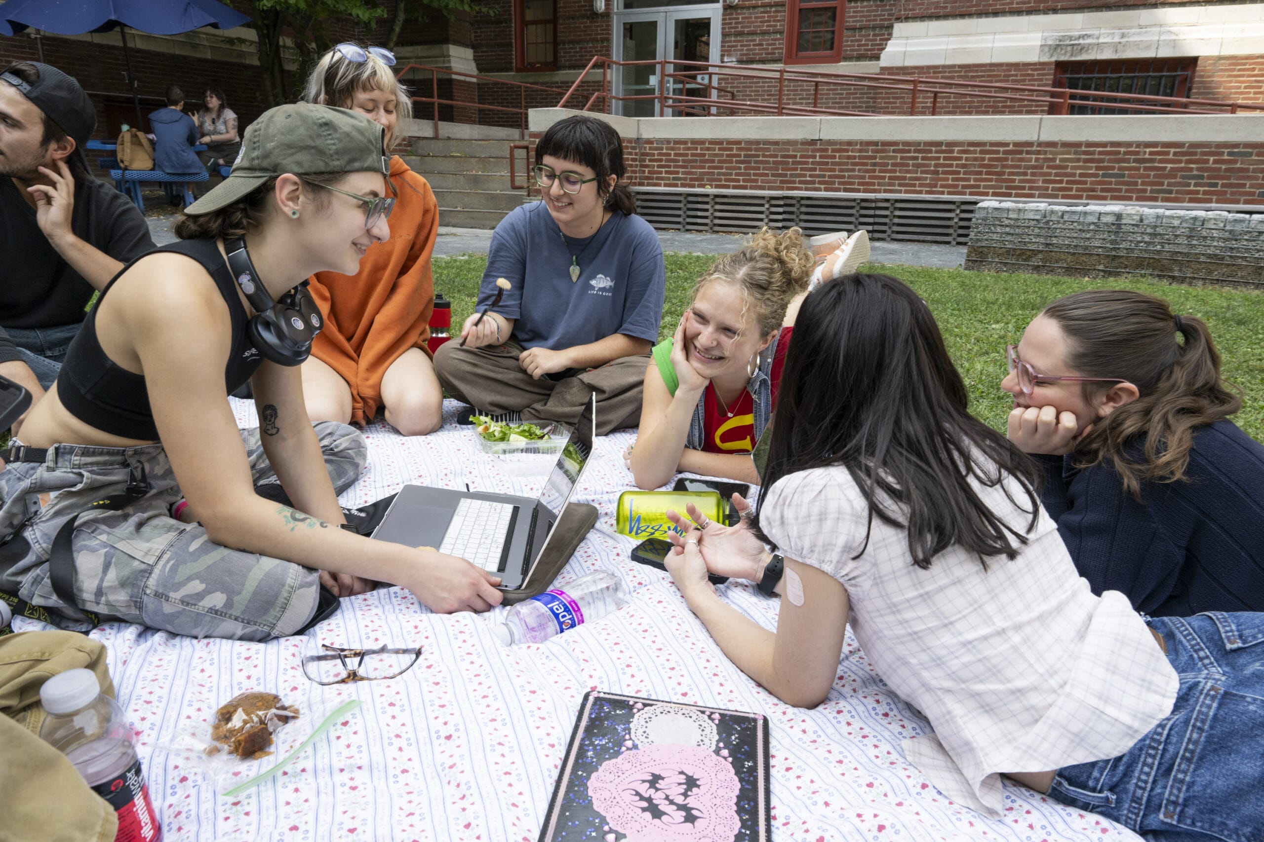 A group of seven people are sitting on a patterned blanket outdoors, smiling and involved in lively conversation. One person with a laptop joins in, while others enjoy snacks and drinks. They are gathered in a grassy area near a brick building.