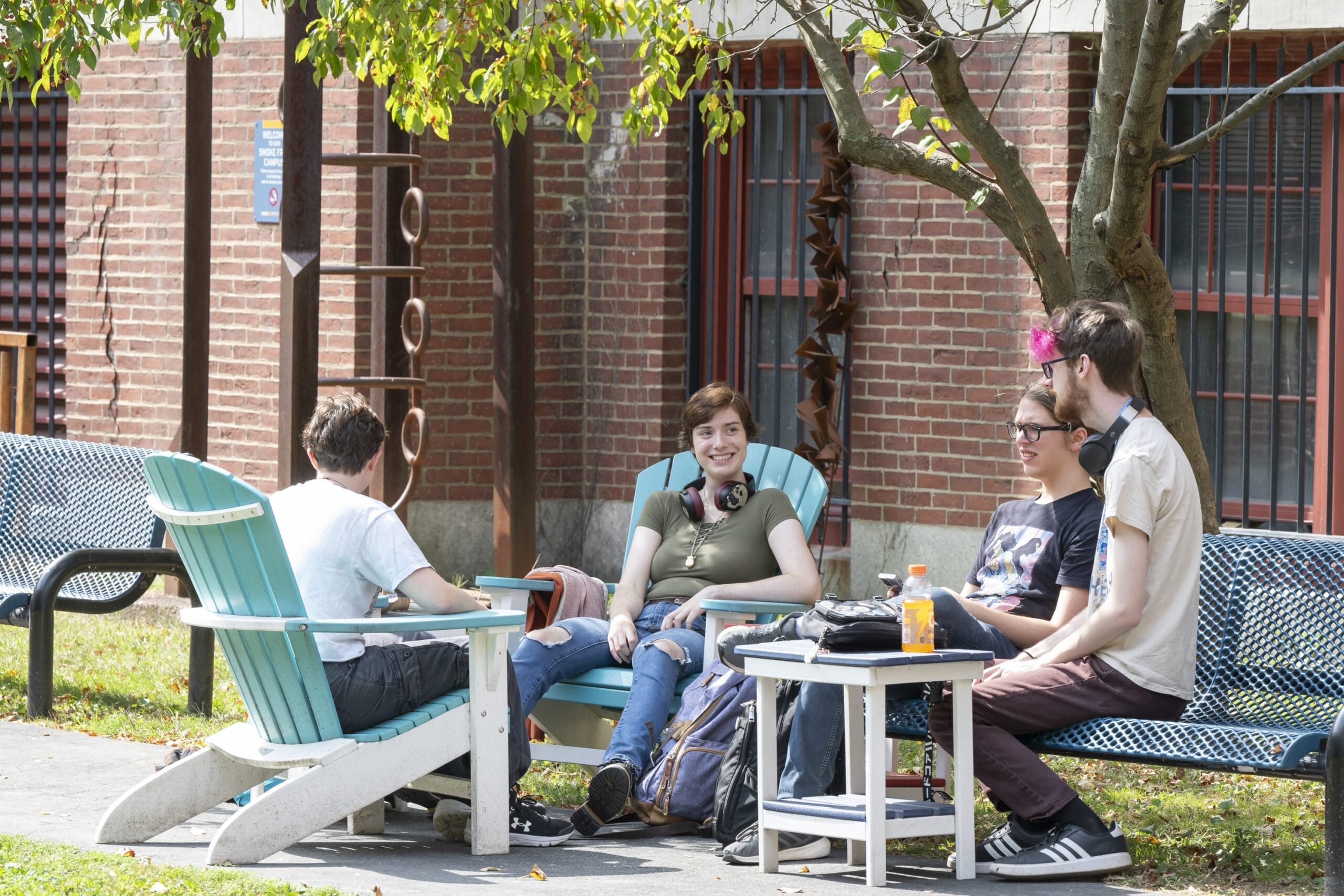 Four people sit and chat in Adirondack chairs, surrounded by backpacks and drinks, under a tree. The inviting outdoor setting, with its brick building and large windows in the background, encourages new friendships. Its the perfect scene to get involved and enjoy the camaraderie.