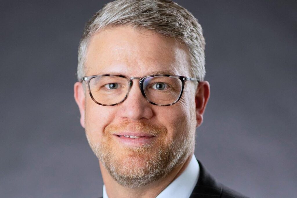 Corey Zink, Vice President of Student Success, stands confidently with short gray hair and glasses. Dressed in a dark suit, light blue shirt, and red striped tie, he poses against a dark background.