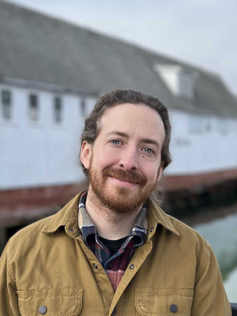 Man with brown hair and beard smiling, wearing a brown jacket and plaid shirt, standing outdoors with a blurred white building in the background.