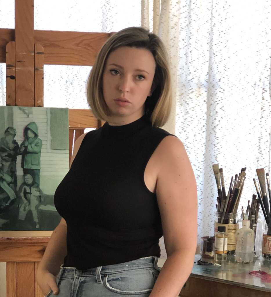 A person with shoulder-length hair, wearing a black sleeveless top and jeans, stands in an art studio. They are near an easel holding a painting of people, with paintbrushes and art supplies on a table nearby.