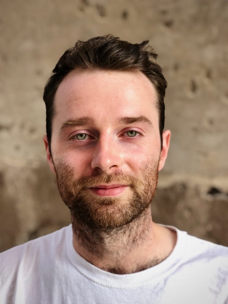 Man with short dark hair and beard wearing a white t-shirt, standing against a textured, neutral-colored background. He has a calm expression.