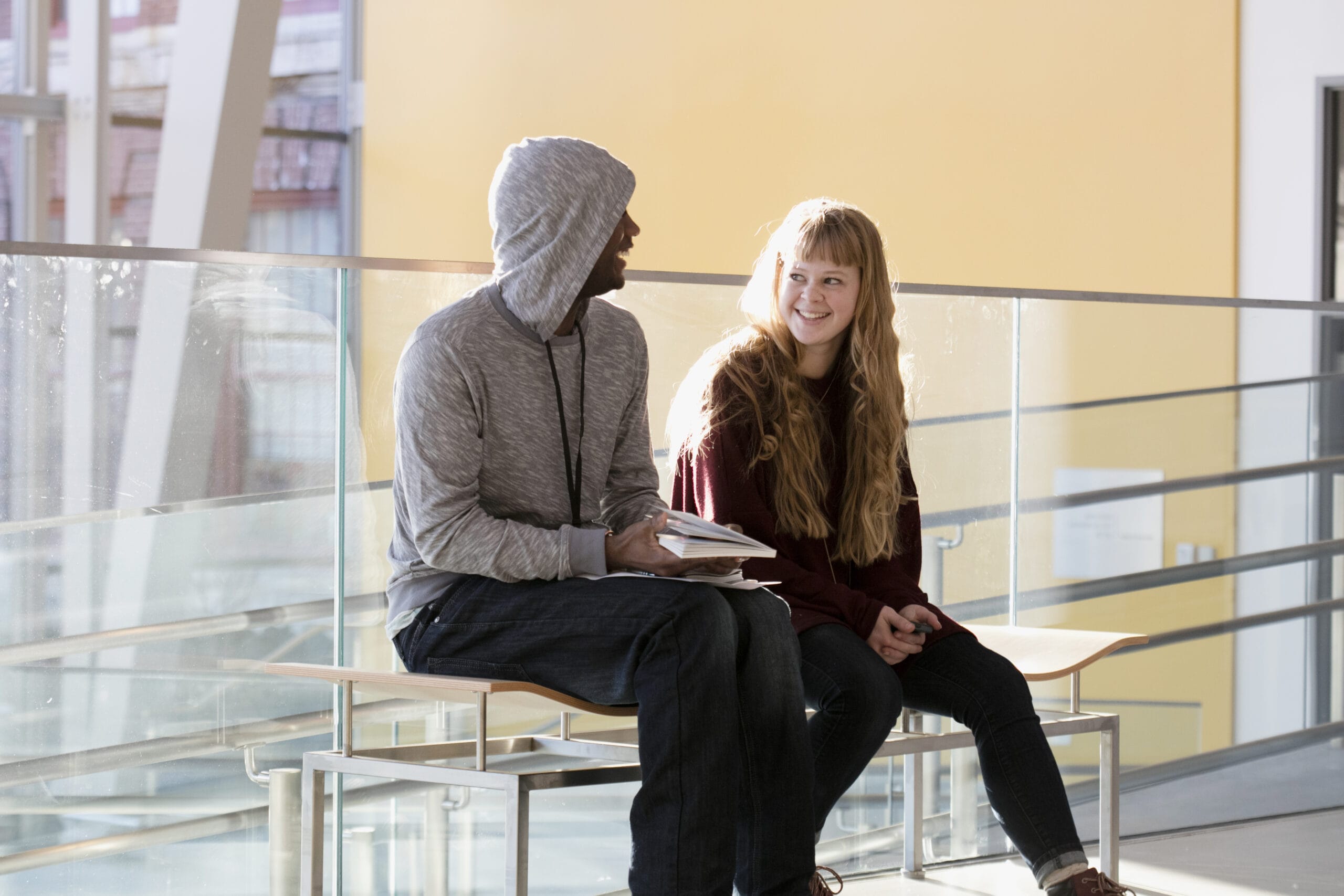 A person in a gray hoodie and jeans sits on a bench next to a person with long hair in a maroon sweater and jeans. Theyre talking about well-being, smiling at each other in a bright, modern interior with large windows.