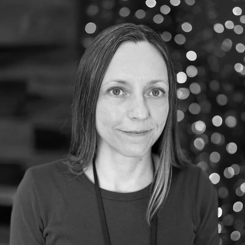 Woman with long straight hair and a serene expression, wearing a dark top, stands in front of a blurred background with circular light patterns, possibly bokeh effects, creating a soft focus backdrop.
