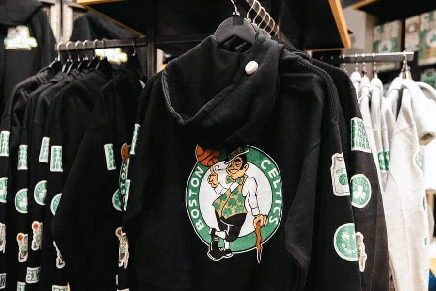 A clothing rack showcases black Boston Celtics hoodies with the iconic leprechaun logo, embodying the spirit of Celtics fashion. Other merchandise, like caps and gray hoodies, completes the scene for fans who embrace this distinctive style.