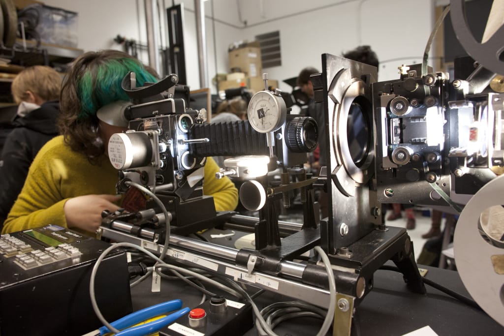 A person with green and black hair expertly manipulates a vintage film editing machine in a bustling workshop. Surrounded by bright lights and various dials, this scene of creativity unfolds with an air of inspiration, observed by others like attentive innovation advisors.