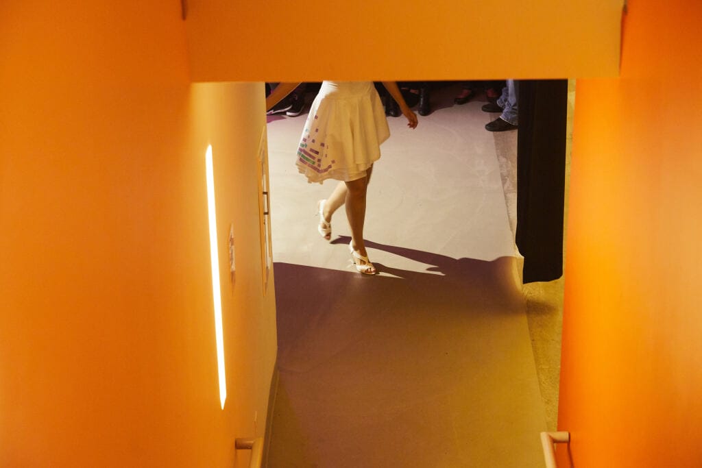 View from a stairway looking down towards a model in a white dress walking off the Fashion Show runway. The orange walls glow softly under the spotlight on the floor, while others gather in the background.