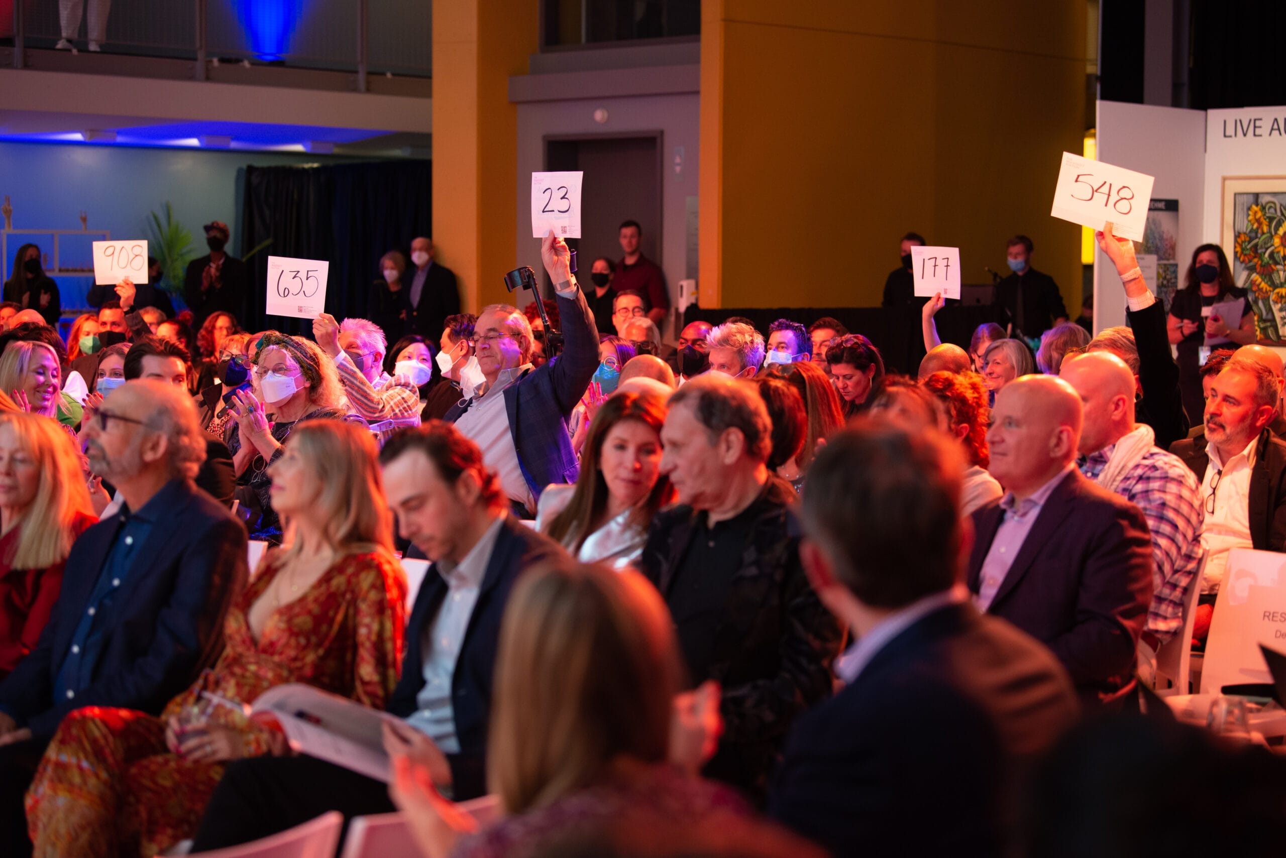 The well-lit auction room buzzes with energy as seated attendees clutch their numbered paddles. Some eagerly raise theirs, displaying numbers like 23 and 548. Amidst the crowd, a colorful artwork peeks from the background, capturing the attention of all engaged bidders.