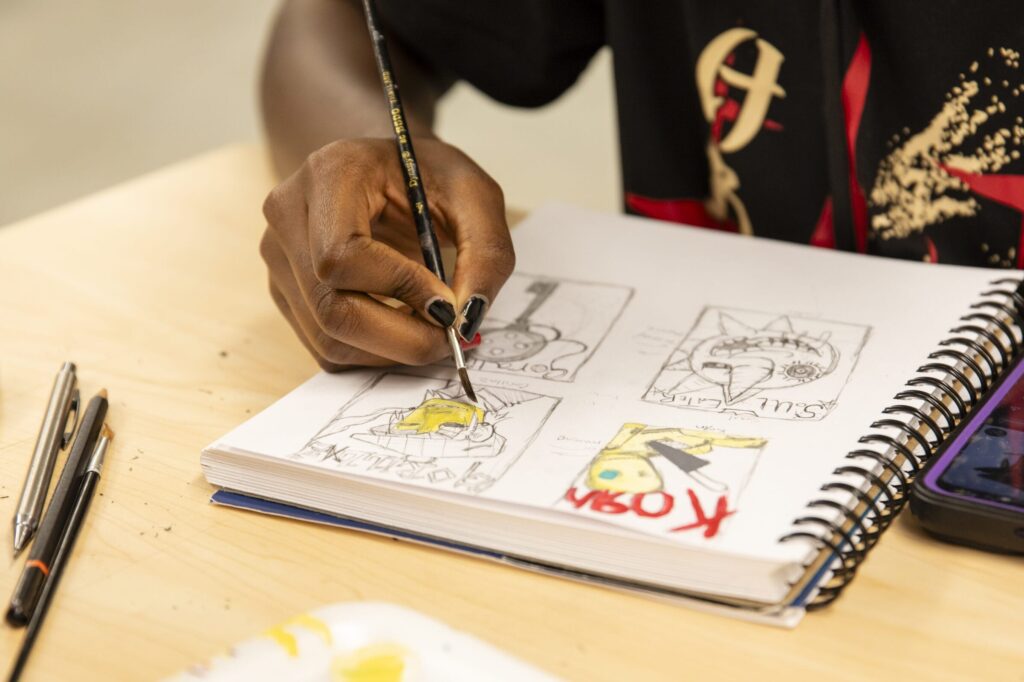 A person is sketching and painting in a spiral-bound notebook, engaging in visual storytelling. The drawings boast various designs, some with numbers and colors like yellow and black. Pens and a phone lie nearby on the table.