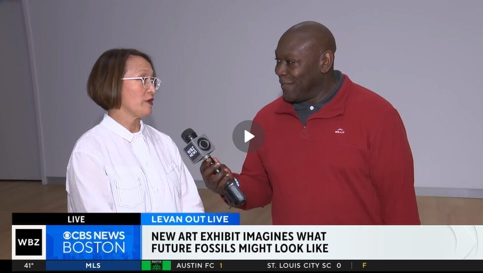 A man in a red sweater interviews a woman in a white shirt for CBS News Boston, discussing the intriguing Boston art exhibit. They stand indoors as the news banner reads, New Art Exhibit Imagines What Future Fossils Might Look Like.