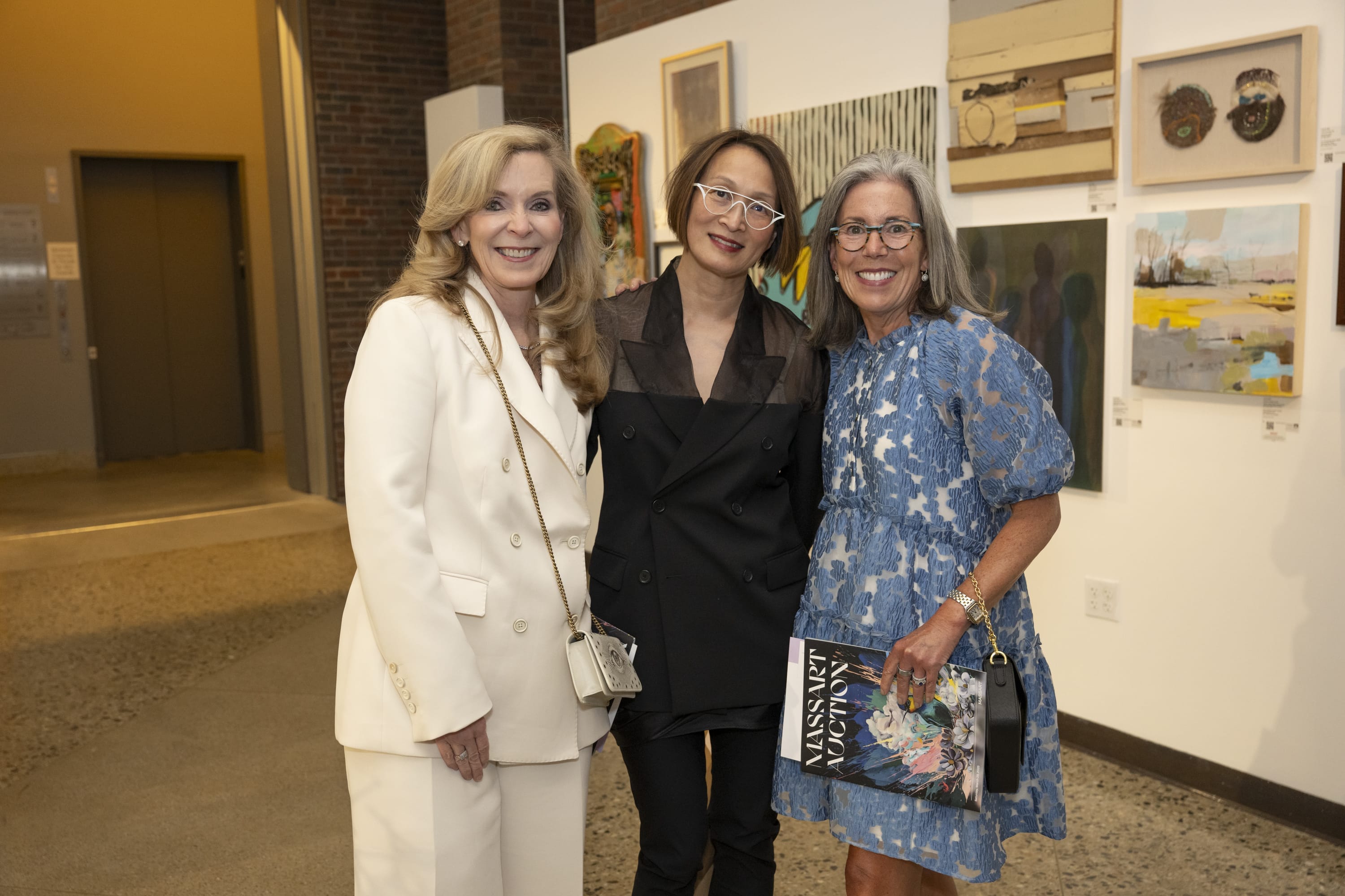 Three women stand together in an art gallery, smiling at the camera. The woman on the left wears a white suit, the middle woman wears black, and the woman on the right wears a blue patterned dress and holds a magazine.