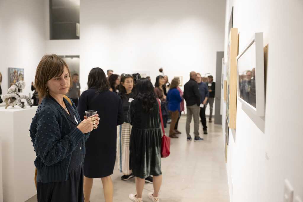 A group of people is gathered in an art gallery, viewing paintings on the walls. A woman in the foreground is holding a drink and looking towards the camera. The setting is modern and minimalistic, with art pieces displayed along the walls and floor.