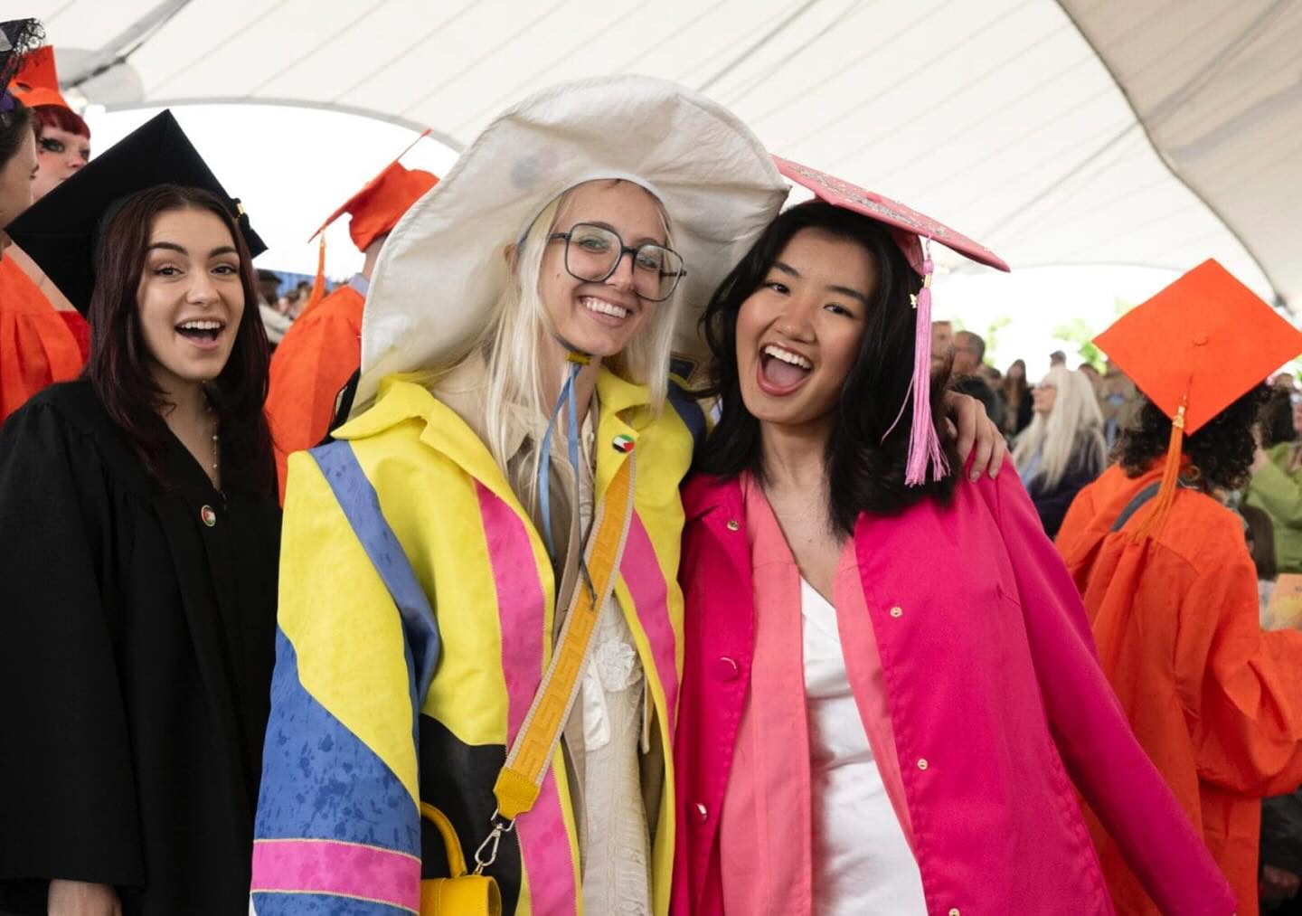 Three graduates smile and pose together at a commencement ceremony. Two wear colorful outfits under their gowns, one in a white hat and glasses, the other in a pink cap and gown. The background shows other graduates in orange caps.