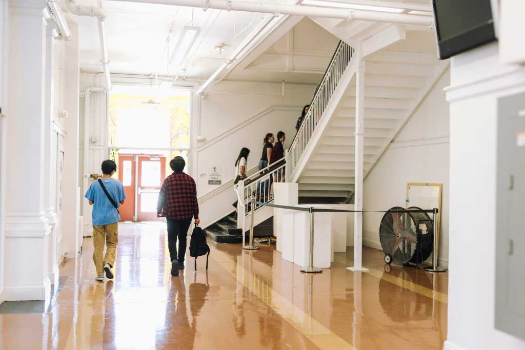 A school hallway buzzes with the anticipation of upcoming events as two students walk away—one in a red plaid shirt, the other in blue. Near the staircase, a cluster of students chat under a large fan. The shiny floor and pristine white walls reflect the excitement of admissions season.