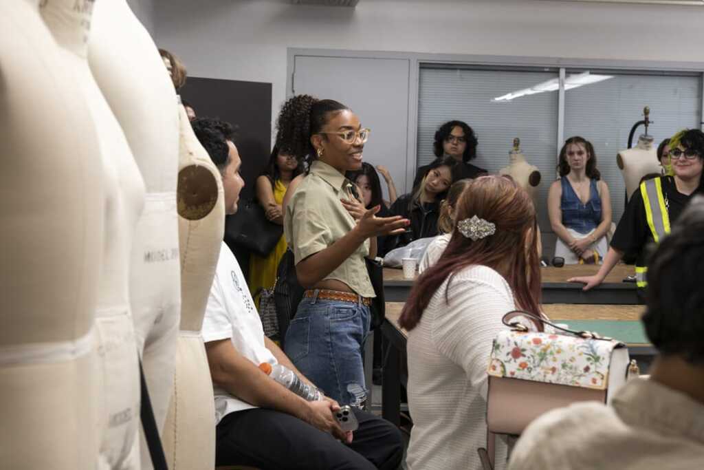 A diverse group of people in a fashion workshop, surrounded by dress forms. A person in a green shirt and glasses speaks enthusiastically while others listen attentively. Fabrics are visible in the background.