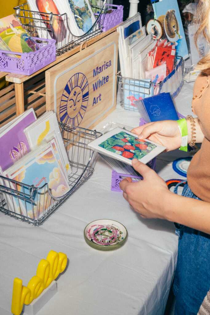 A person looks at a colorful print at an art market table displaying various prints, cards, and ceramics. A wooden sign reads Marisa White Art among baskets and arranged artwork.