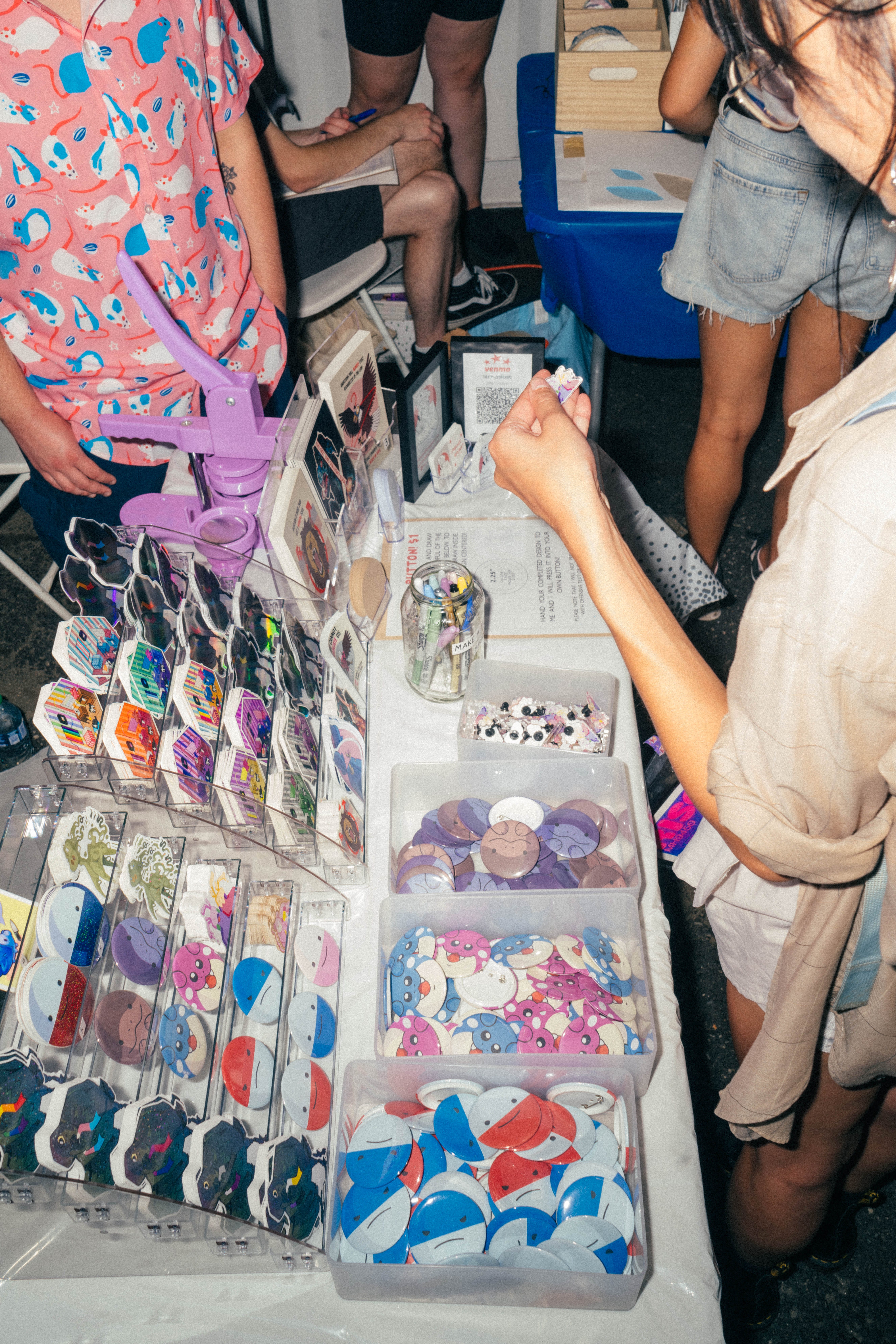 A person browses a colorful table display of buttons and pins at a market stall, with others standing nearby. The table is covered with trays and racks holding various cartoon and illustrated pins.