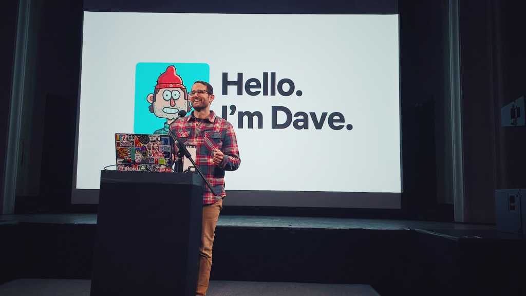 A man stands at a podium covered in stickers, giving a presentation. Behind him, a screen displays a cartoon avatar and the text: Hello. Im Dave Schlafman—an artist known for his work with MassArt and Netflix.