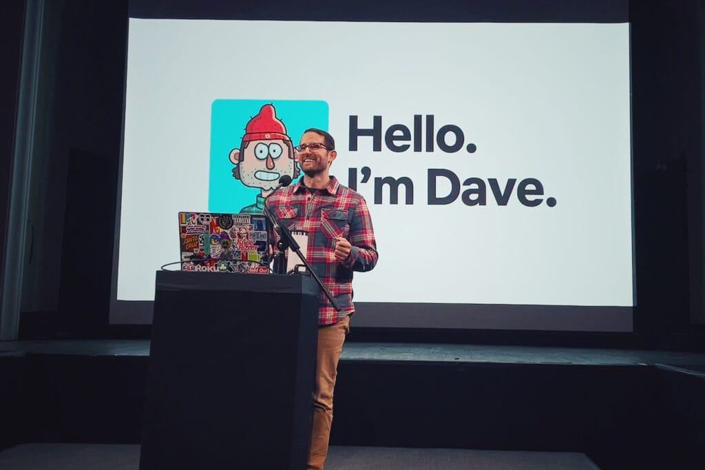 A man stands at a podium covered in stickers, giving a presentation. Behind him, a screen displays a cartoon avatar and the text: Hello. Im Dave Schlafman—an artist known for his work with MassArt and Netflix.