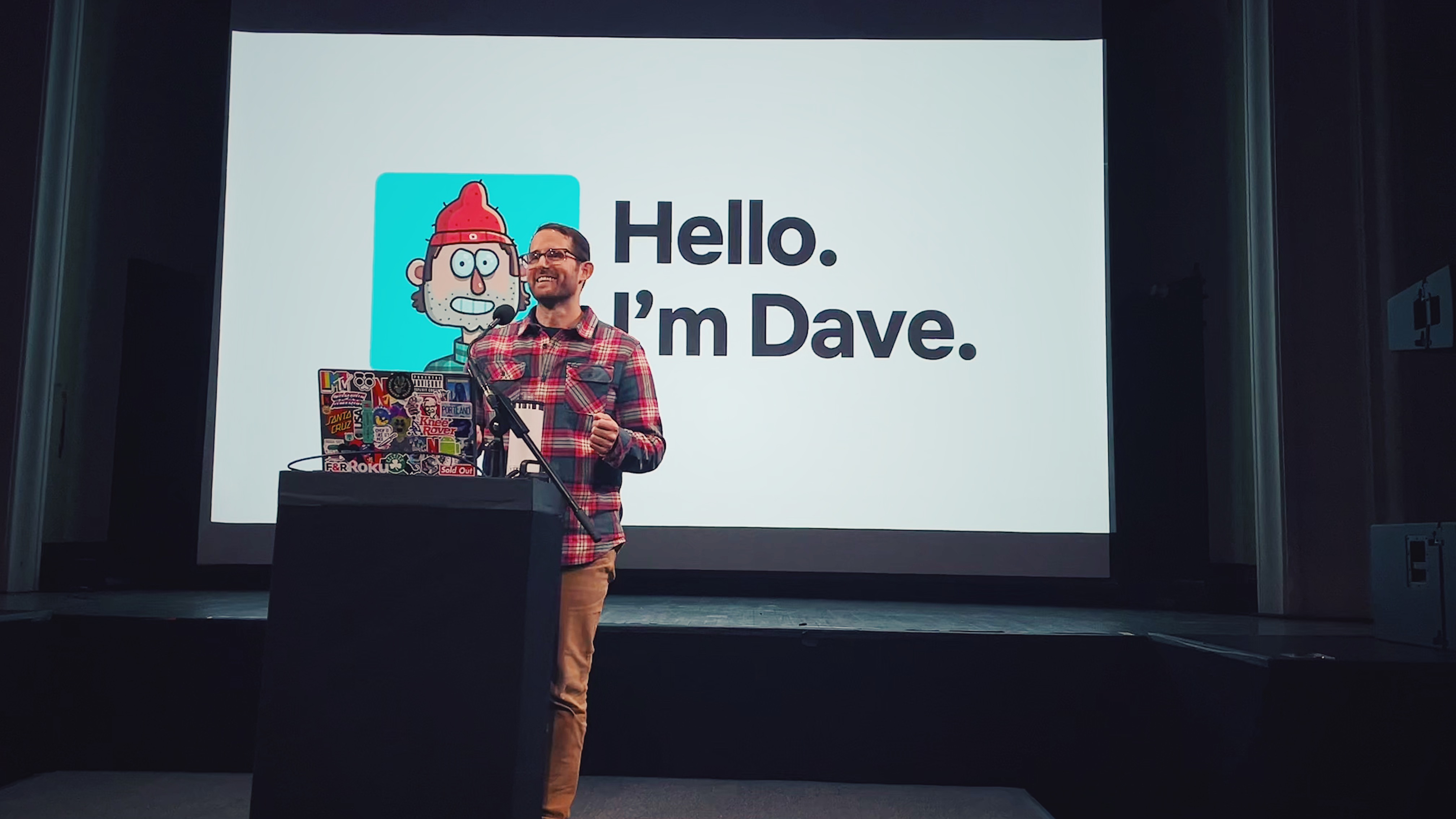 A man stands at a podium covered in stickers, giving a presentation. Behind him, a screen displays a cartoon avatar and the text: Hello. Im Dave Schlafman—an artist known for his work with MassArt and Netflix.