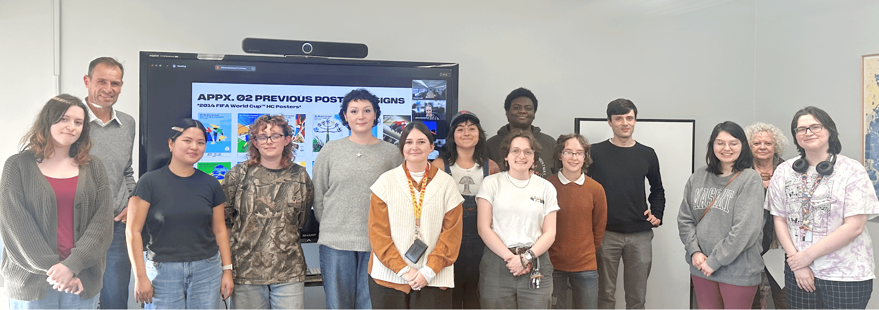 A group of fifteen people stand in a room at MassArt, smiling at the camera. Behind them is a large screen displaying colorful images and text. The room is well-lit, with a painting visible on the wall to the right.