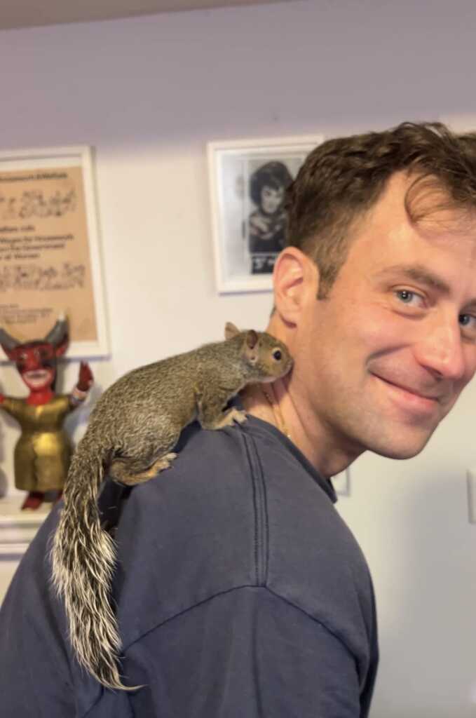 A smiling man in a blue shirt stands indoors with a gray squirrel perched on his shoulder. Framed pictures and a red devil figurine are visible on the wall in the background.
