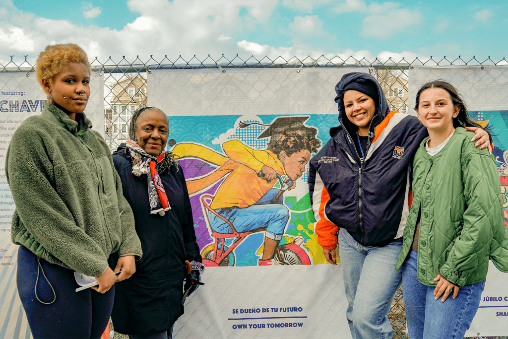 Four women stand smiling in front of vibrant street art on a chain-link fence. The mural features a child in a graduation cap and superhero cape, riding a tricycle, adding charm to these unique chain link galleries. The sky is partly cloudy.