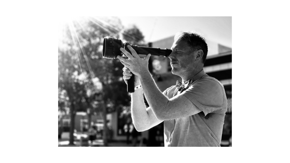 A man in a short-sleeved shirt stands outdoors, holding a video camera up to his eye and filming. Sunlight streams in from the top left, creating a bright, high-contrast scene with trees in the background—capturing moments like a MassArt student.