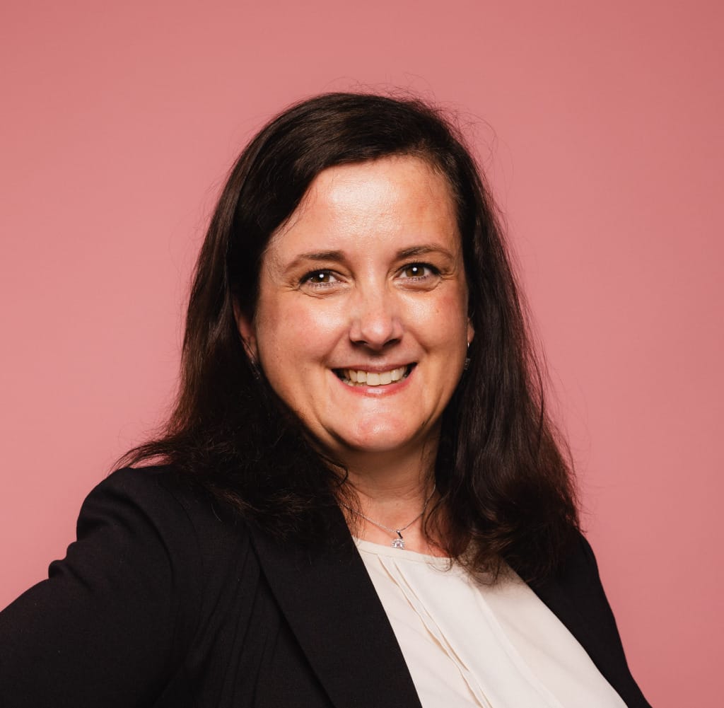 A person with shoulder-length dark hair smiles at the camera. They are wearing a black blazer over a light blouse. The background is a solid pink color.