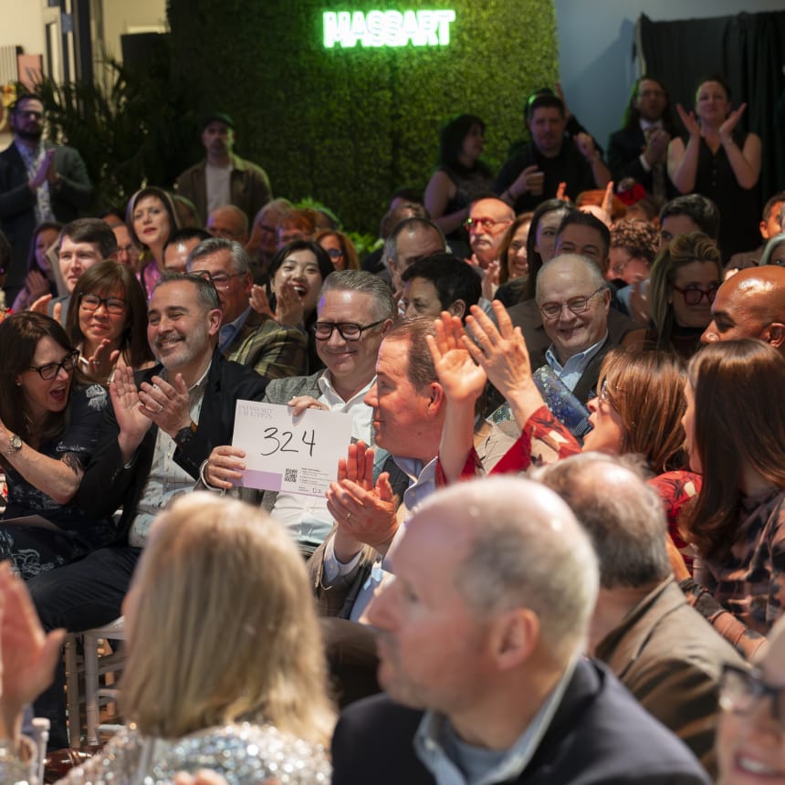 A lively crowd seated at an indoor event applauds enthusiastically. A man near the center holds up a sign with the number 324. The setting is well-lit with a mix of casual and formal attire. A neon Hasart sign glows in the background.
