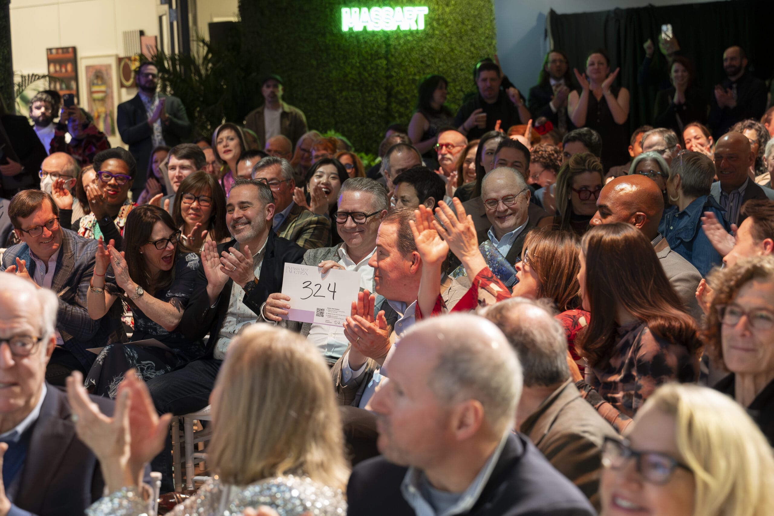 A lively crowd seated at an indoor event applauds enthusiastically. A man near the center holds up a sign with the number 324. The setting is well-lit with a mix of casual and formal attire. A neon Hasart sign glows in the background.