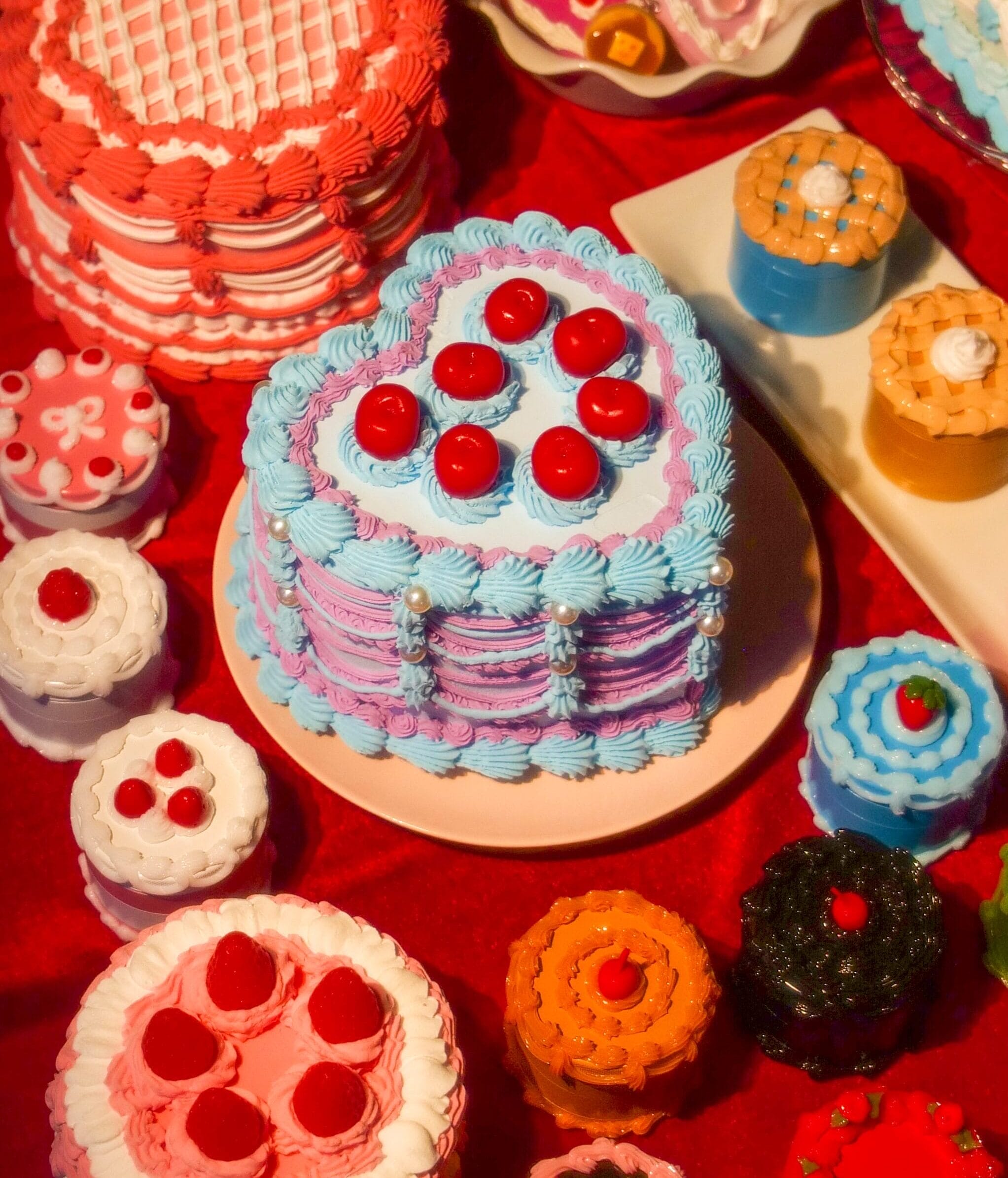 A variety of colorful, intricately decorated cakes and pastries are arranged on a red surface, with a heart-shaped blue and purple cake topped with cherries at the center—showcasing creativity reminiscent of MassArt.
