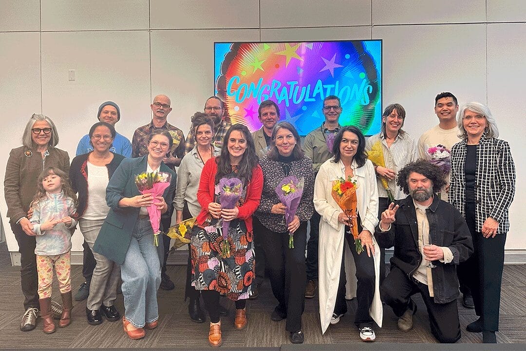 A group of people stand smiling indoors, some holding bouquets, in front of a colorful screen that says Congratulations. The group appears to be celebrating a faculty promotion or tenure achievement at MassArt.