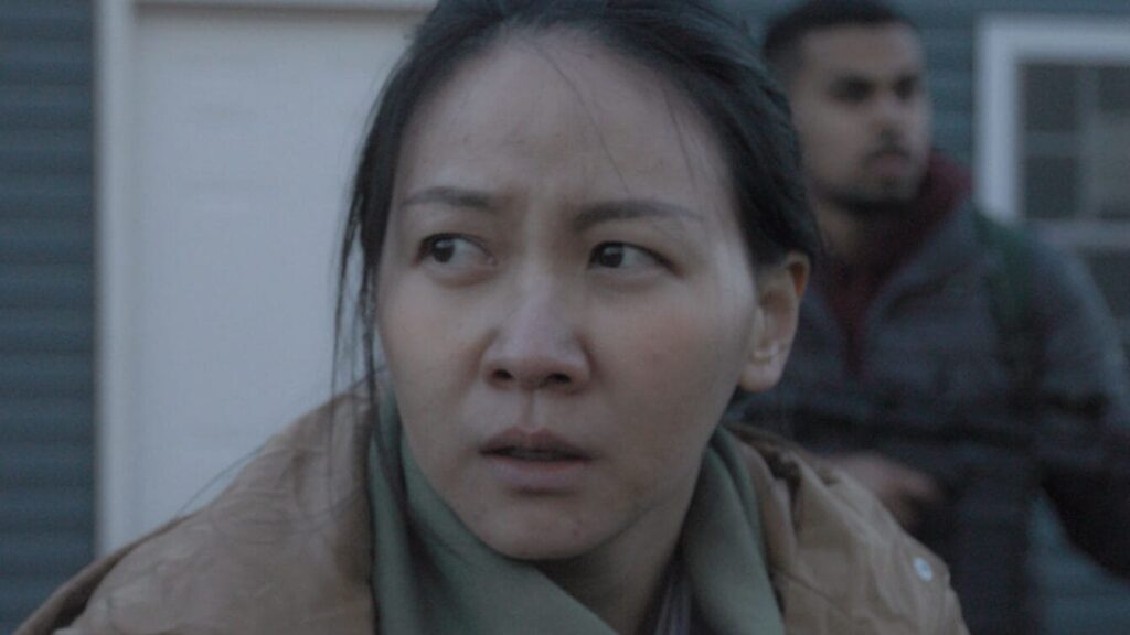 A woman with a worried expression looks to the side, standing outside near a blue house; a man holding a rifle appears blurred in the background at the Boston Student Showcase presented by MassArt.