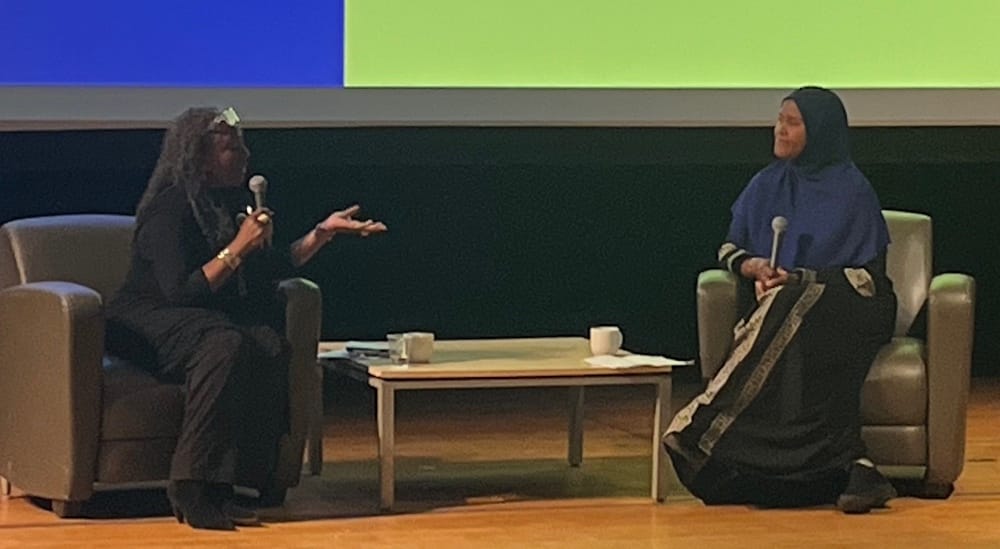 Ekua Holmes and Autumn Allen sit in armchairs on stage, engaged in lively conversation. Separated by a small table with mugs, they discuss Connected Legacies against a vibrant, colorful backdrop.